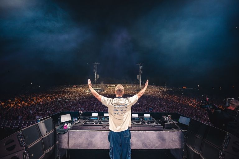 DJ Calvin Harris seen from behind with arms raised overlooking a massive outdoor festival crowd from the main stage decks during a live set.